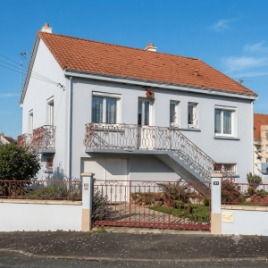 Vue extérieure d'une maison de ville de style Nantaise typique avec un enduit gris clair, un toit en tuiles rouges et un escalier extérieur avec des balustrades en fer forgé rouge. La maison dispose d'un garage au rez-de-chaussée et d'un portail assorti sur une rue pavée.